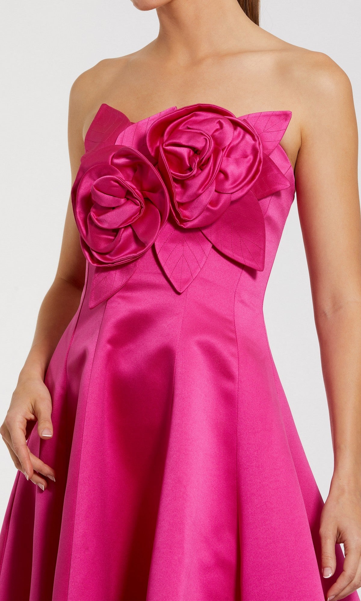 A woman wears the Mac Duggal 2303 High-Low Formal Dress, a strapless satin evening gown in bright pink with a high-low hem and bold 3D floral and leaf embellishments on the bodice.