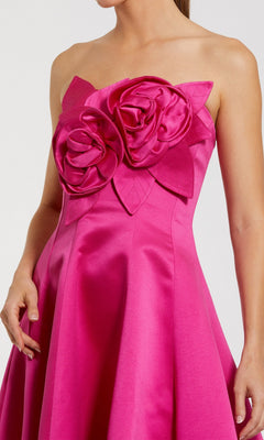 A woman wears the Mac Duggal 2303 High-Low Formal Dress, a strapless satin evening gown in bright pink with a high-low hem and bold 3D floral and leaf embellishments on the bodice.