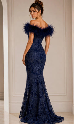 A woman stands facing away in a neutral-toned room, wearing the Jovani 23030 long formal dress, which features an off-the-shoulder neckline and beaded floral lace accented with feather trim.