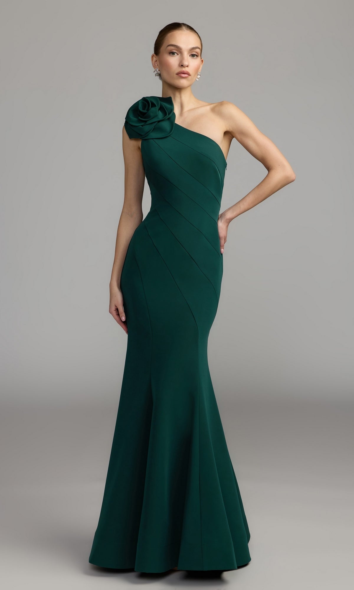 An elegant woman models the Mac Duggal 2304 Flower One-Shoulder Formal Dress in emerald, featuring a fitted silhouette and a striking large fabric flower accent on the single shoulder, against a plain grey background.