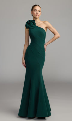 An elegant woman models the Mac Duggal 2304 Flower One-Shoulder Formal Dress in emerald, featuring a fitted silhouette and a striking large fabric flower accent on the single shoulder, against a plain grey background.