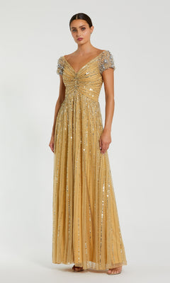 A woman models the Mac Duggal 23042, a short sleeve gold formal dress with sequins and a fitted bodice, against a plain white background, highlighting this elegant evening gown.