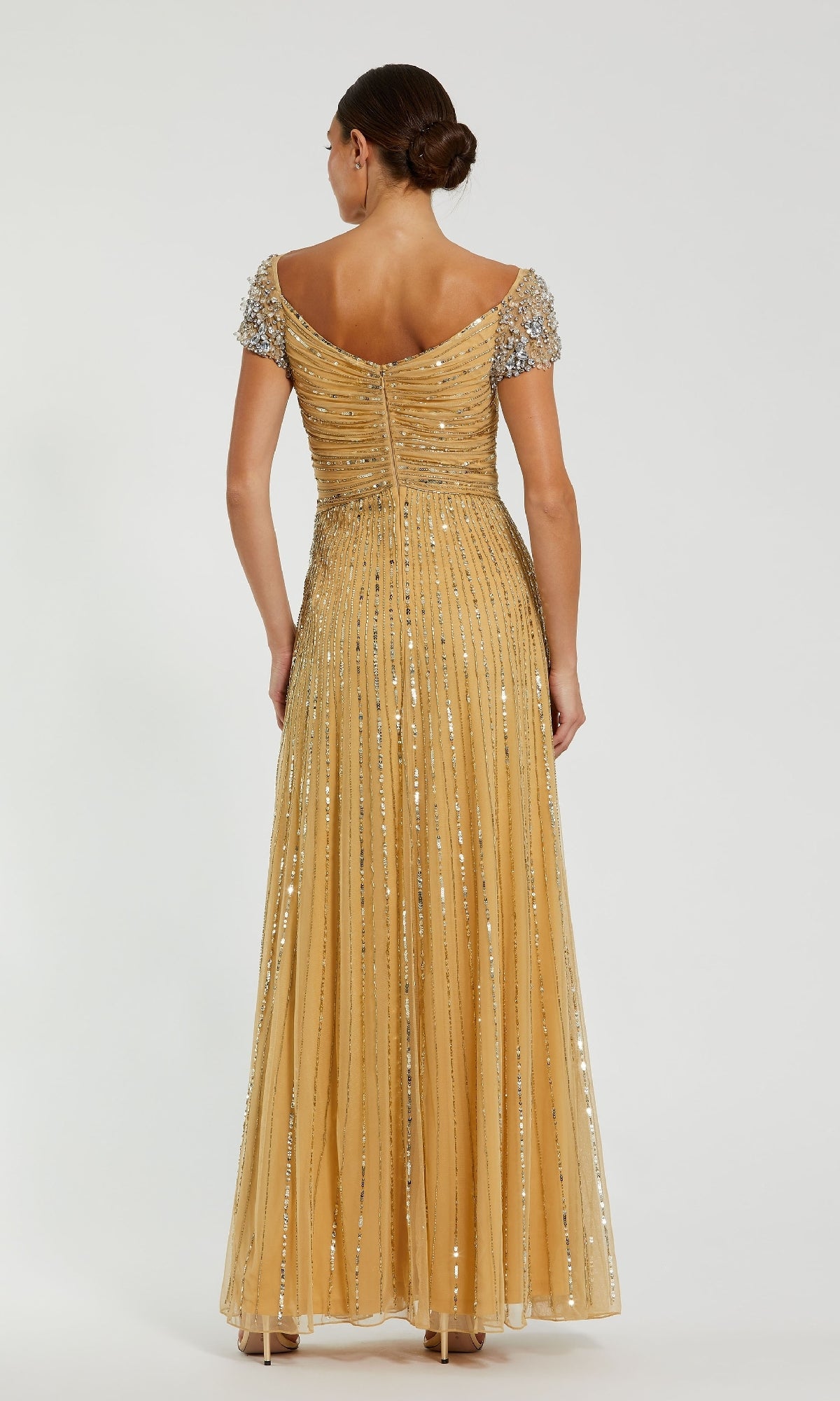 A woman faces away, wearing the Mac Duggal 23042 short sleeve gold formal dress with sequin detailing and a fitted bodice. Her hair is styled in a low bun.