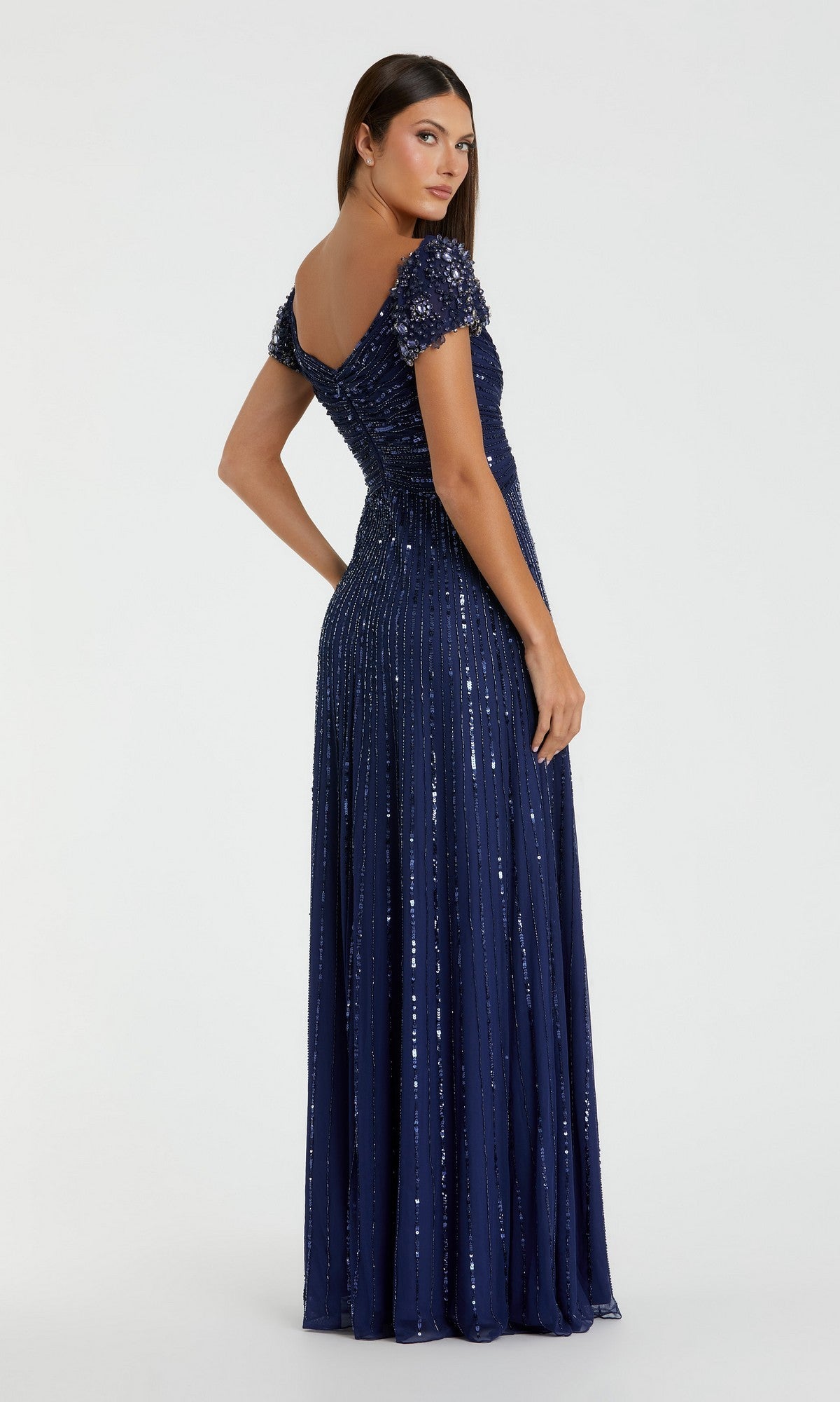 A woman stands facing away, wearing the Mac Duggal 23042 short sleeve gold formal dress with a fitted bodice and sparkling sequined detailing, set against a plain light background.