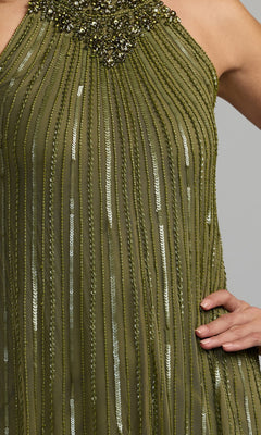 A close-up of the Mac Duggal 23043 long formal dress, featuring a sleeveless olive green design with vertical beaded stripes and an embellished neckline. A person’s hand is resting on their hip.