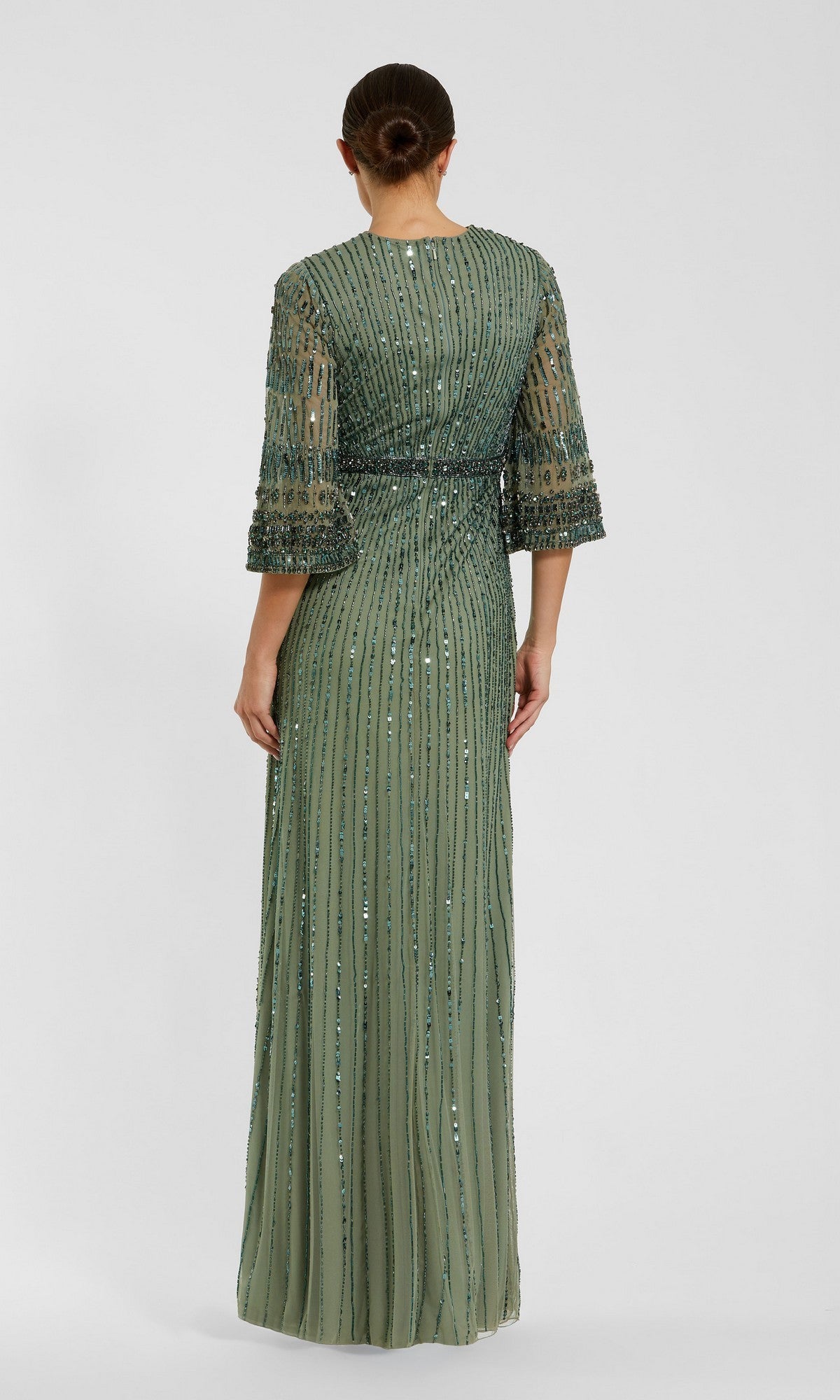 A person stands facing away, wearing the Mac Duggal 23045 long formal dress in sage green, featuring floor-length beaded vertical stripes and three-quarter sleeves, with their hair styled in a low bun.