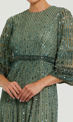A person wearing the Mac Duggal 23045 long formal dress—a green, sequined gown with sheer three-quarter sleeves and vertical bead embellishments—stands with hands clasped.