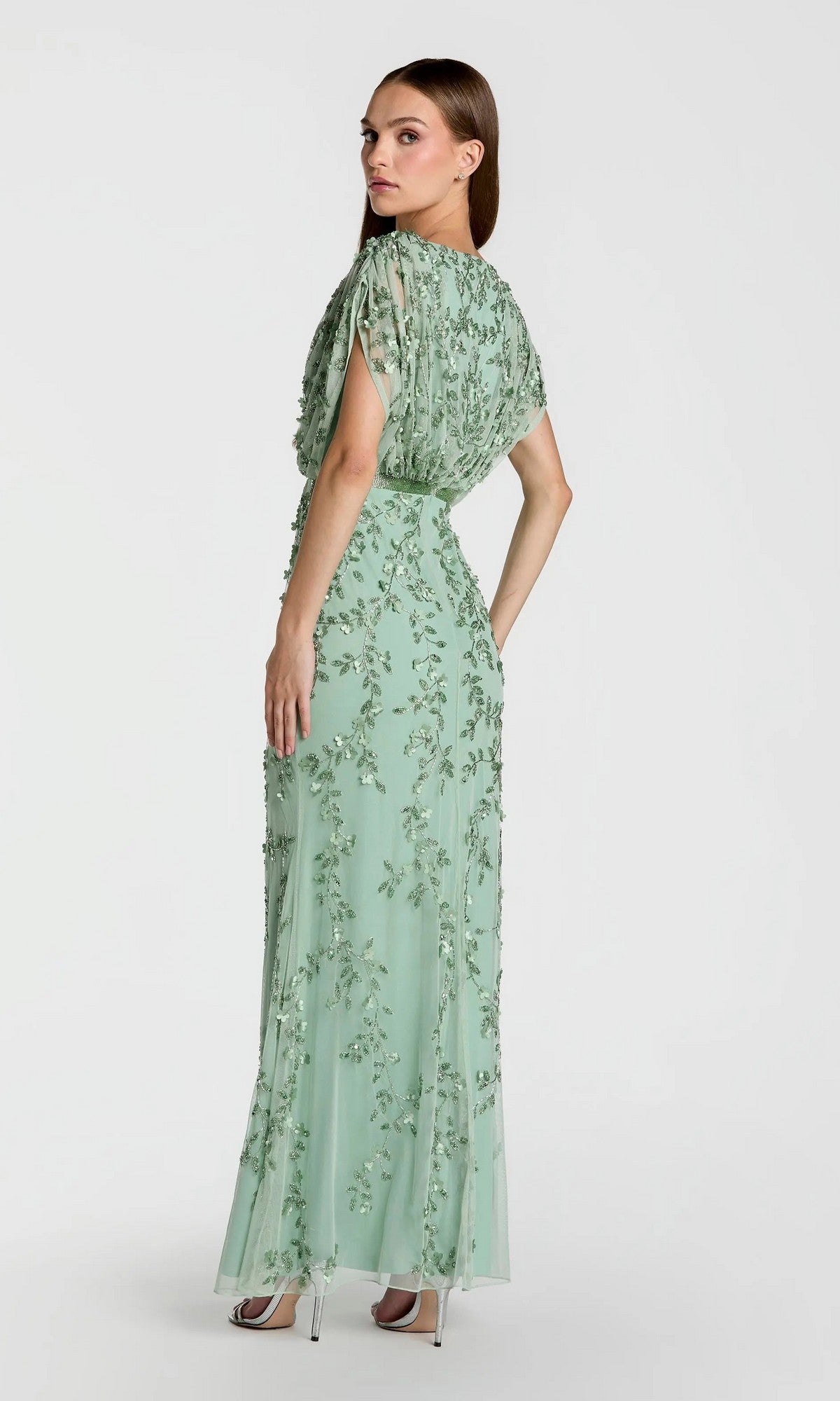 A woman is wearing the Mac Duggal 23067 long formal dress, a floor-length light green beaded gown with intricate floral embroidery. She stands in heels, facing away and looking over her shoulder against a plain background.