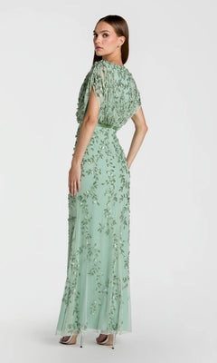 A woman is wearing the Mac Duggal 23067 long formal dress, a floor-length light green beaded gown with intricate floral embroidery. She stands in heels, facing away and looking over her shoulder against a plain background.