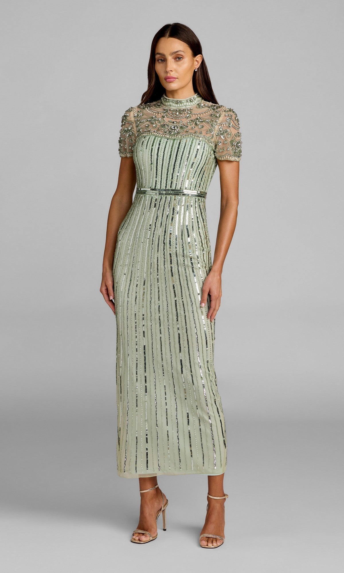 A woman stands against a plain gray background, wearing the Mac Duggal 23075 Long Formal Dress in light sage—a floor-length, sequined gown with short sleeves and a sheer beaded bodice—paired with strappy high heels.
