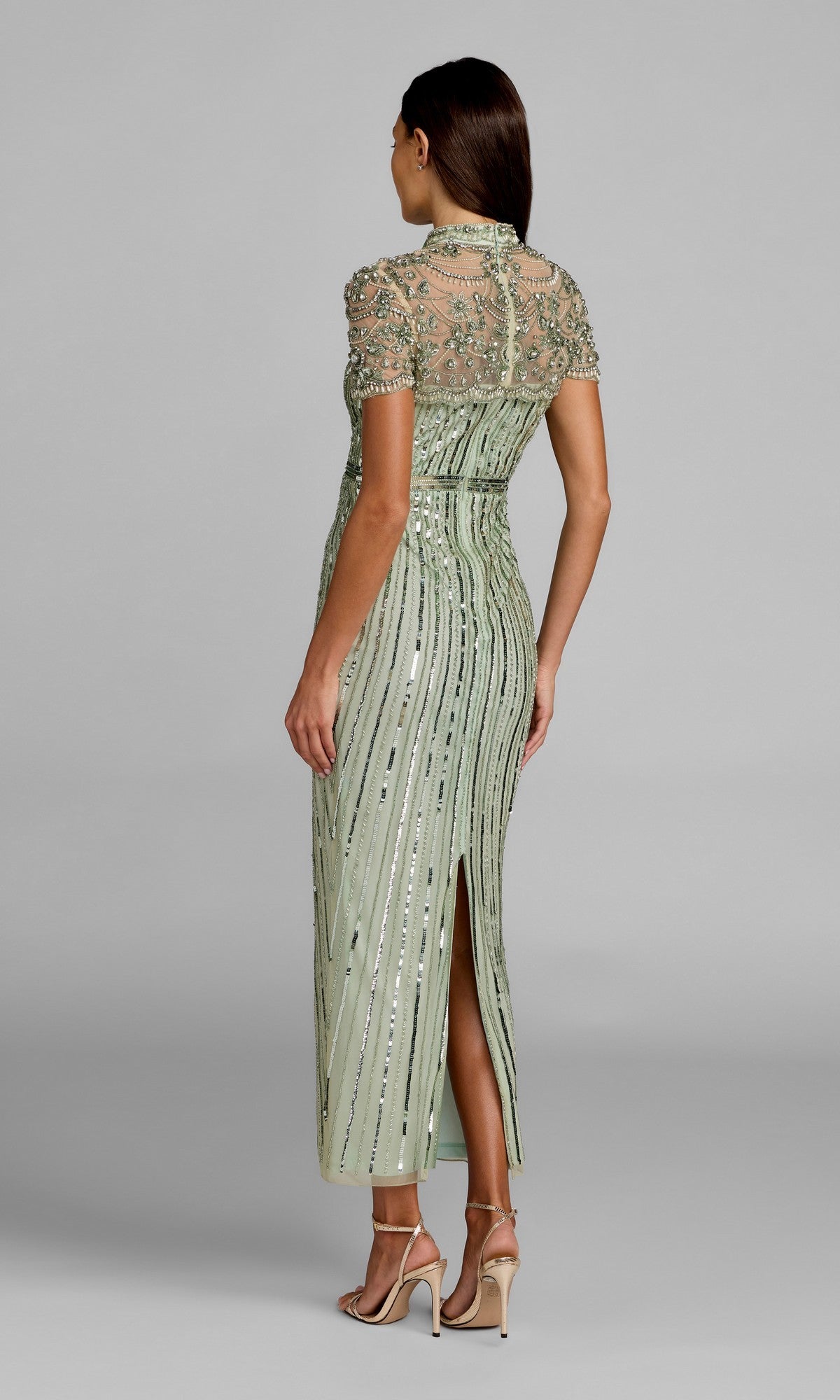 A woman stands facing away in the Mac Duggal 23075 long formal dress, a light green, beaded design with short sleeves, a sheer embellished top, and a back slit; she wears high-heeled sandals.