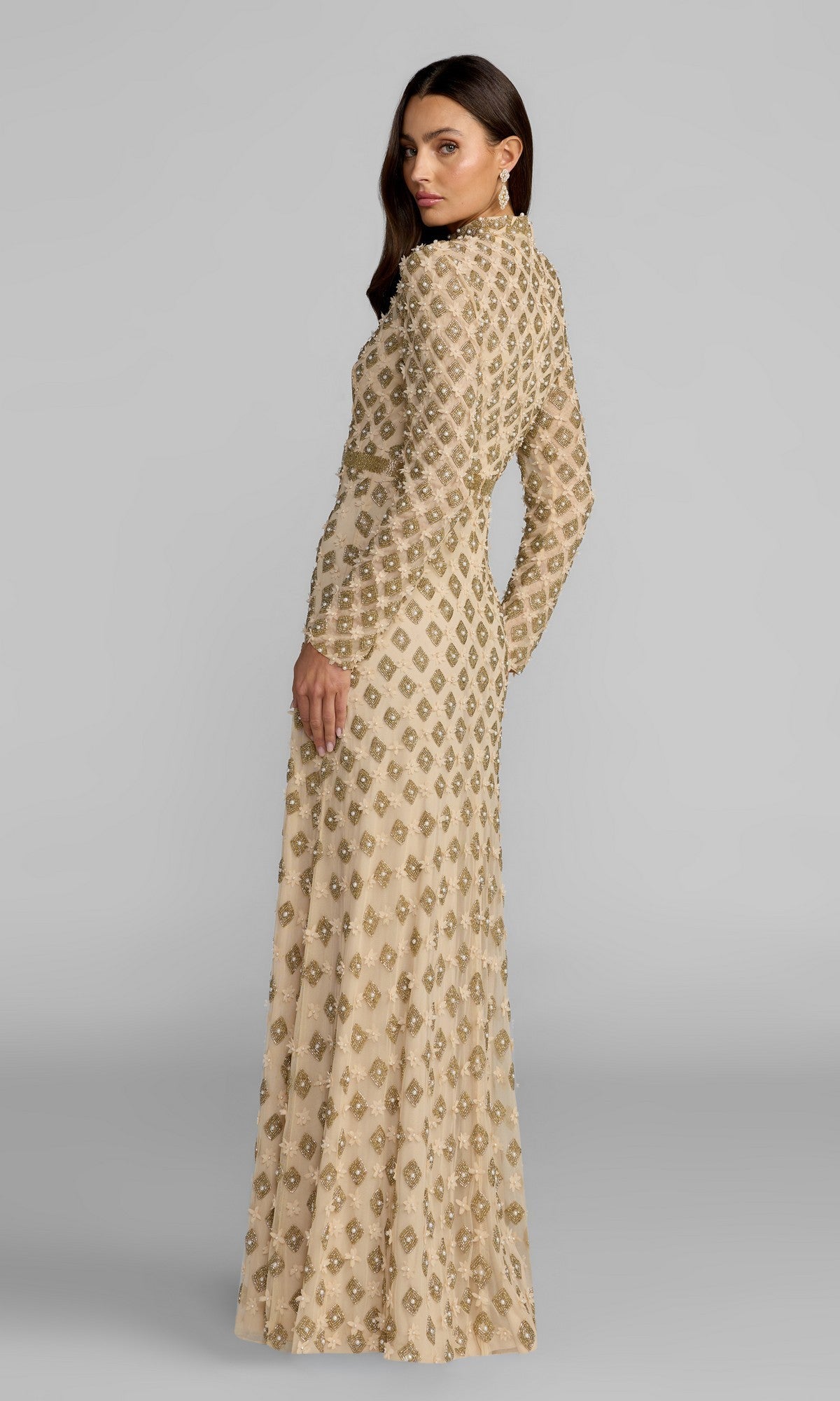 A woman wearing the Mac Duggal 23087 long formal dress, a beige gown with gold embellishments and long sleeves, stands facing away while turning her head over her shoulder.