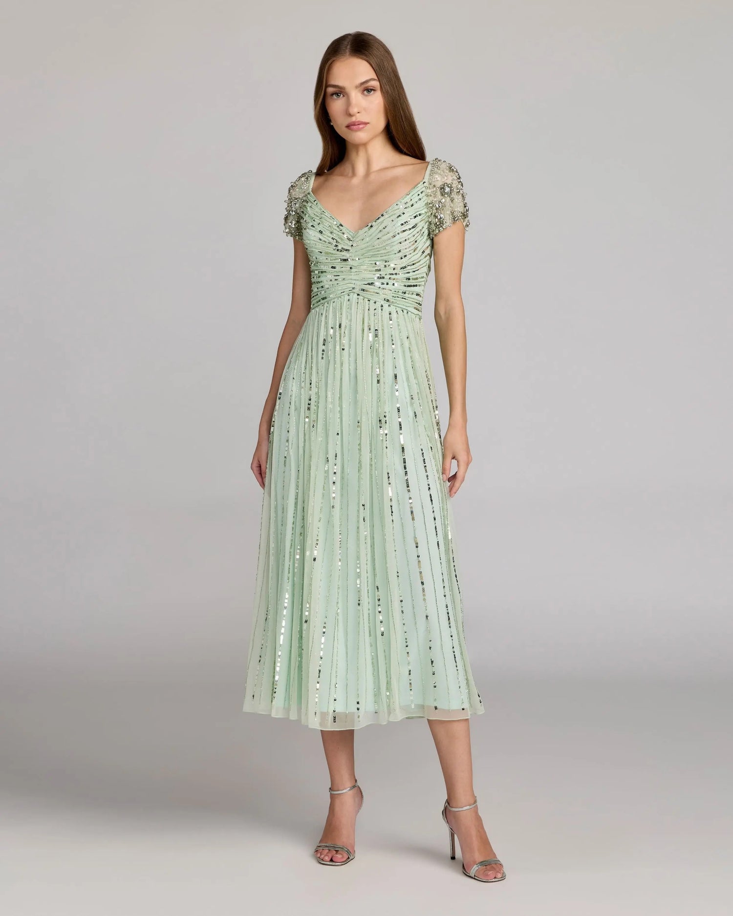 A woman stands wearing the Midi Party Dress: Mac Duggal 23098 in sage, which features beaded short sleeves, a ruched bodice, and a pleated, A-line skirt adorned with sequins. She has long, straight hair and is accessorized with silver heels.