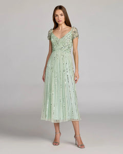 A woman stands wearing the Midi Party Dress: Mac Duggal 23098 in sage, which features beaded short sleeves, a ruched bodice, and a pleated, A-line skirt adorned with sequins. She has long, straight hair and is accessorized with silver heels.