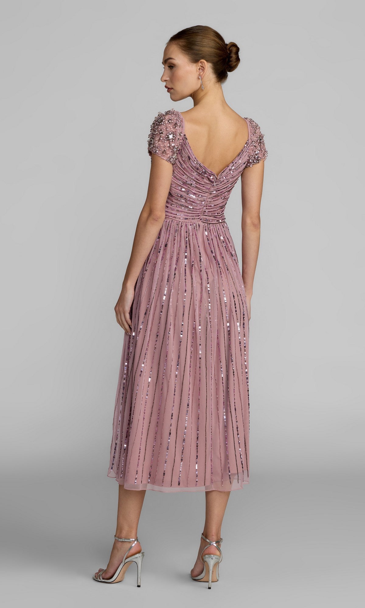 A woman stands facing away, wearing the Mac Duggal 23098 midi party dress in mauve, featuring shimmering sequins, beaded short sleeves, a V-back, and an elegant pleated A-line skirt. Her hair is styled in a neat bun and she completes the look with silver high-heeled sandals.