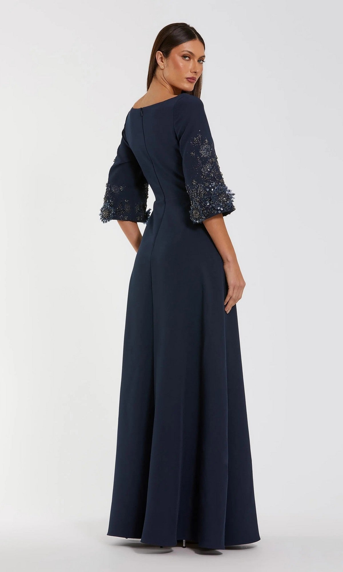 A woman in the Mac Duggal 2311 Long Formal Dress, a floor-length navy crepe gown with three-quarter sleeves, lace, and beaded details, stands against a plain light background, facing slightly away.