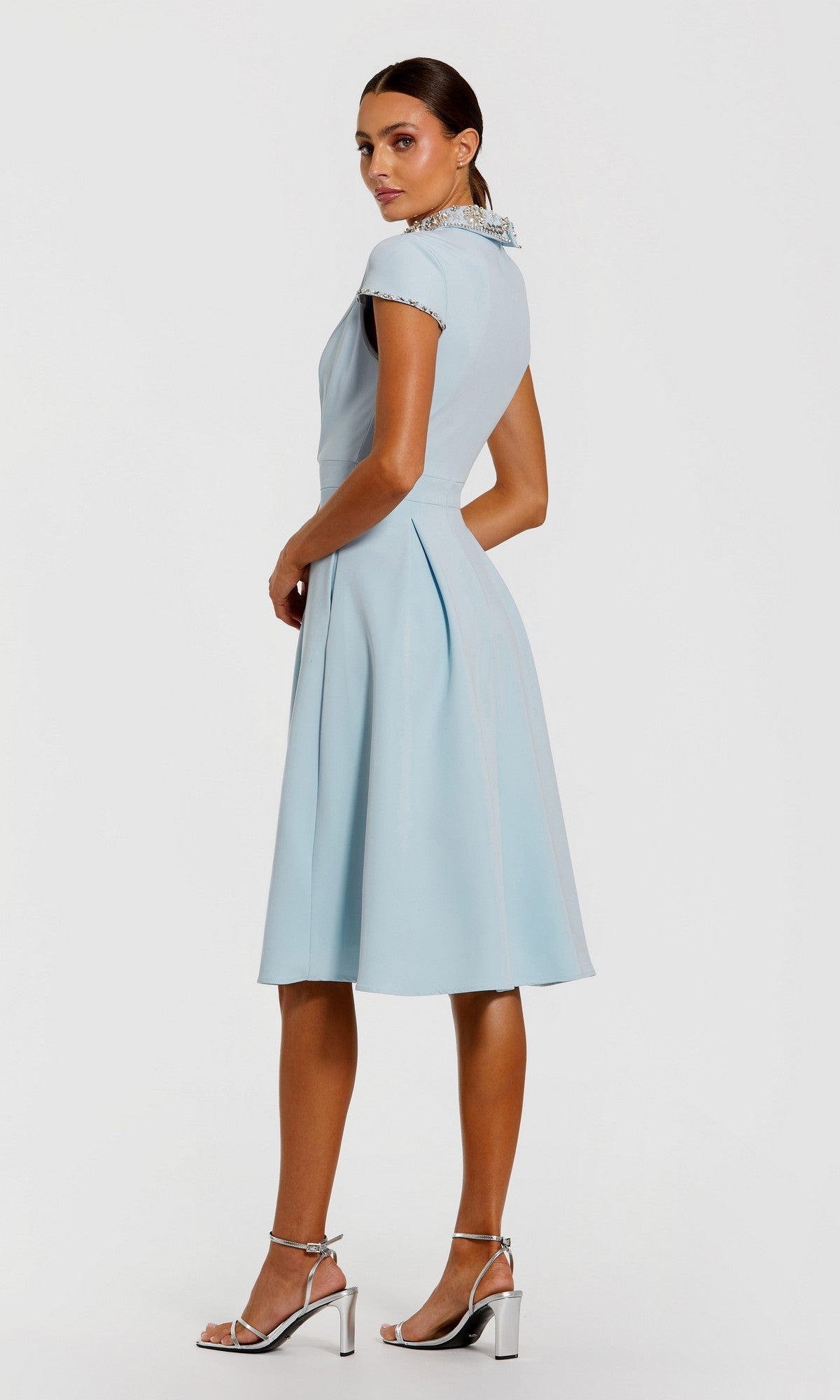 A woman in profile wears the Mac Duggal 2313 Midi Wedding Guest Dress—light blue with cap sleeves, an a-line skirt, and a crystal-embellished collar—paired with silver high-heeled sandals, set against a plain white background.