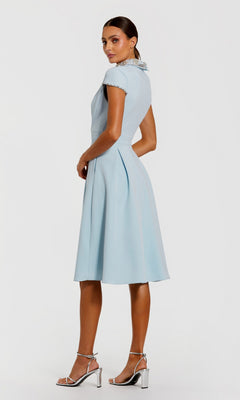 A woman in profile wears the Mac Duggal 2313 Midi Wedding Guest Dress—light blue with cap sleeves, an a-line skirt, and a crystal-embellished collar—paired with silver high-heeled sandals, set against a plain white background.