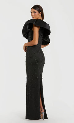 Dressed in the Mac Duggal 2315 long black formal gown featuring dramatic ruffled shoulder detailing and subtle embellishments, the woman poses against a plain white background, accessorized with silver high-heeled sandals.