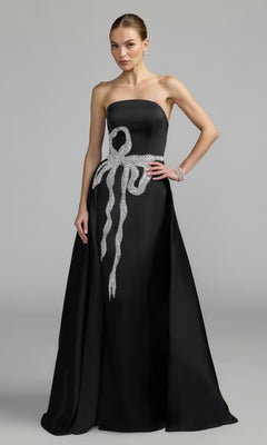 Woman wearing a strapless long formal dress by Mac Duggal (Style 2317), featuring a floor-length black and white gown with a silver jeweled bow design on the bodice and hip, standing against a plain gray background.