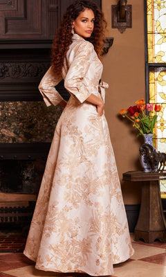 Wearing the Jovani 23178 Long Formal Dress in cream and gold brocade, a woman with long curly hair stands indoors beside a table adorned with flowers, with a stained glass window in the background.
