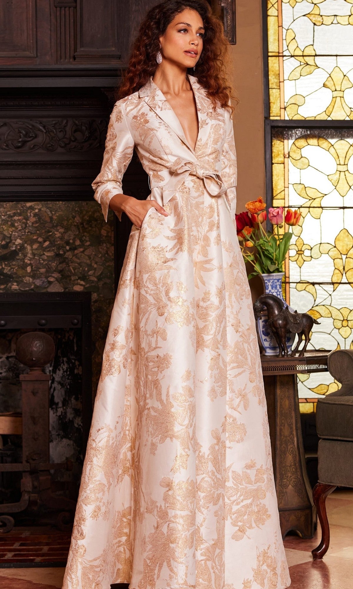 A woman stands indoors beside a stained glass window and a table with a vase of flowers, wearing the Jovani 23178—a long, cream and gold floral-patterned metallic Mikado formal dress featuring a deep neckline and pockets.