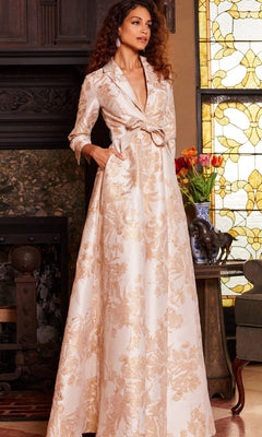 A woman stands indoors beside a stained glass window and a table with a vase of flowers, wearing the Jovani 23178—a long, cream and gold floral-patterned metallic Mikado formal dress featuring a deep neckline and pockets.