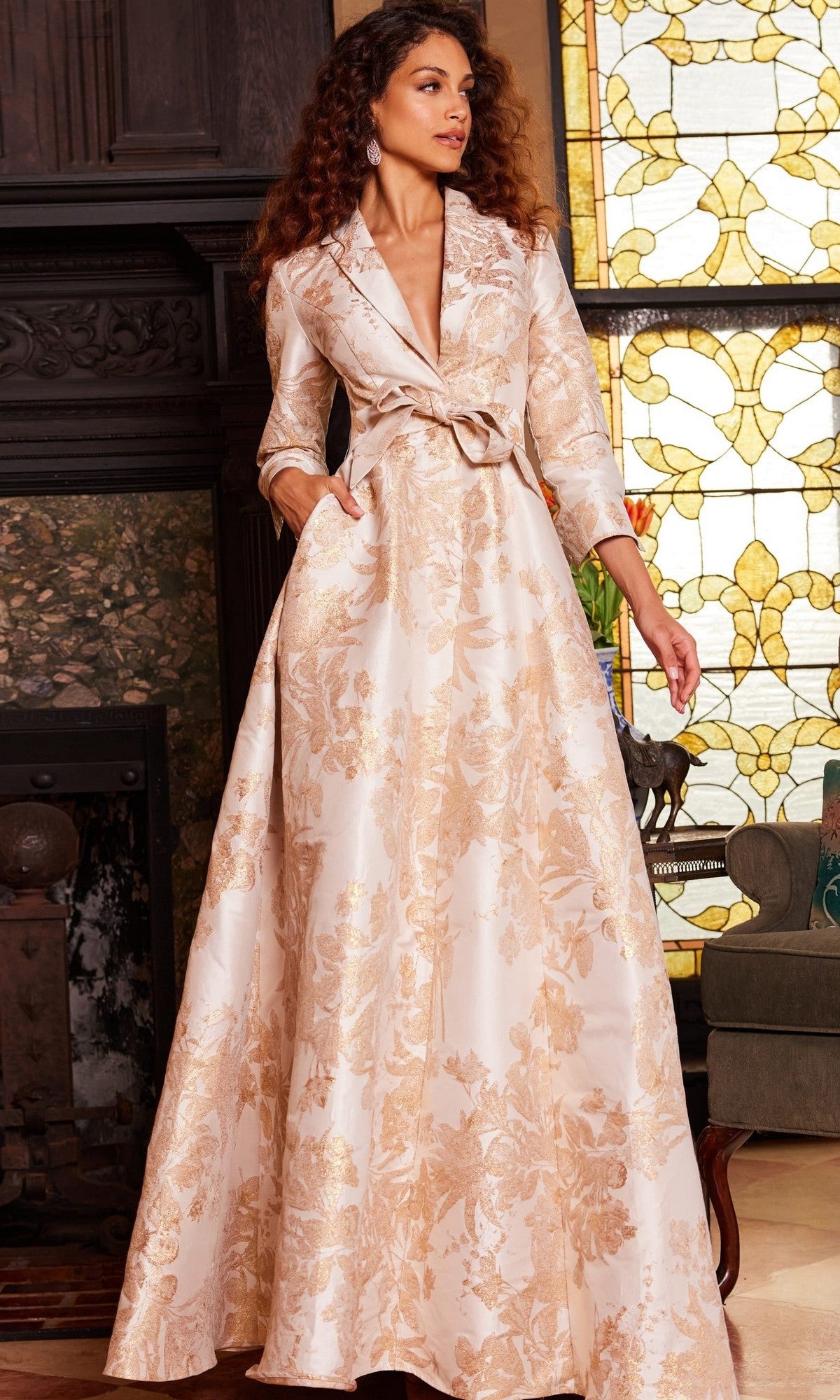 A woman wearing the Jovani 23178, a long formal designer dress in cream and gold floral brocade with a shawl collar, stands indoors near stained glass windows and a fireplace.