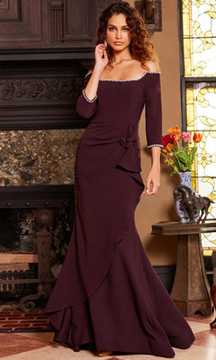A woman in a long, off-the-shoulder Jovani Long Formal Dress with Bead Trim 23190 in dark burgundy stands indoors by a fireplace and a table decorated with flowers and a horse statue.