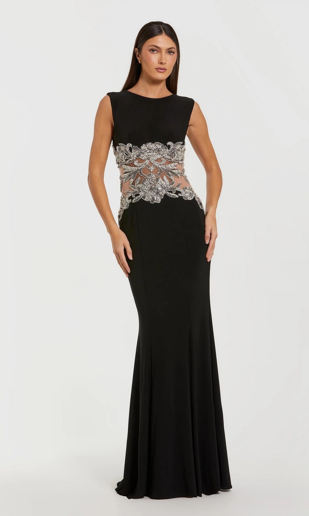 A woman stands against a white background in the Mac Duggal 2324 long formal black dress, which has a sleeveless floor-length design and a beaded illusion waist with silver floral embroidery.