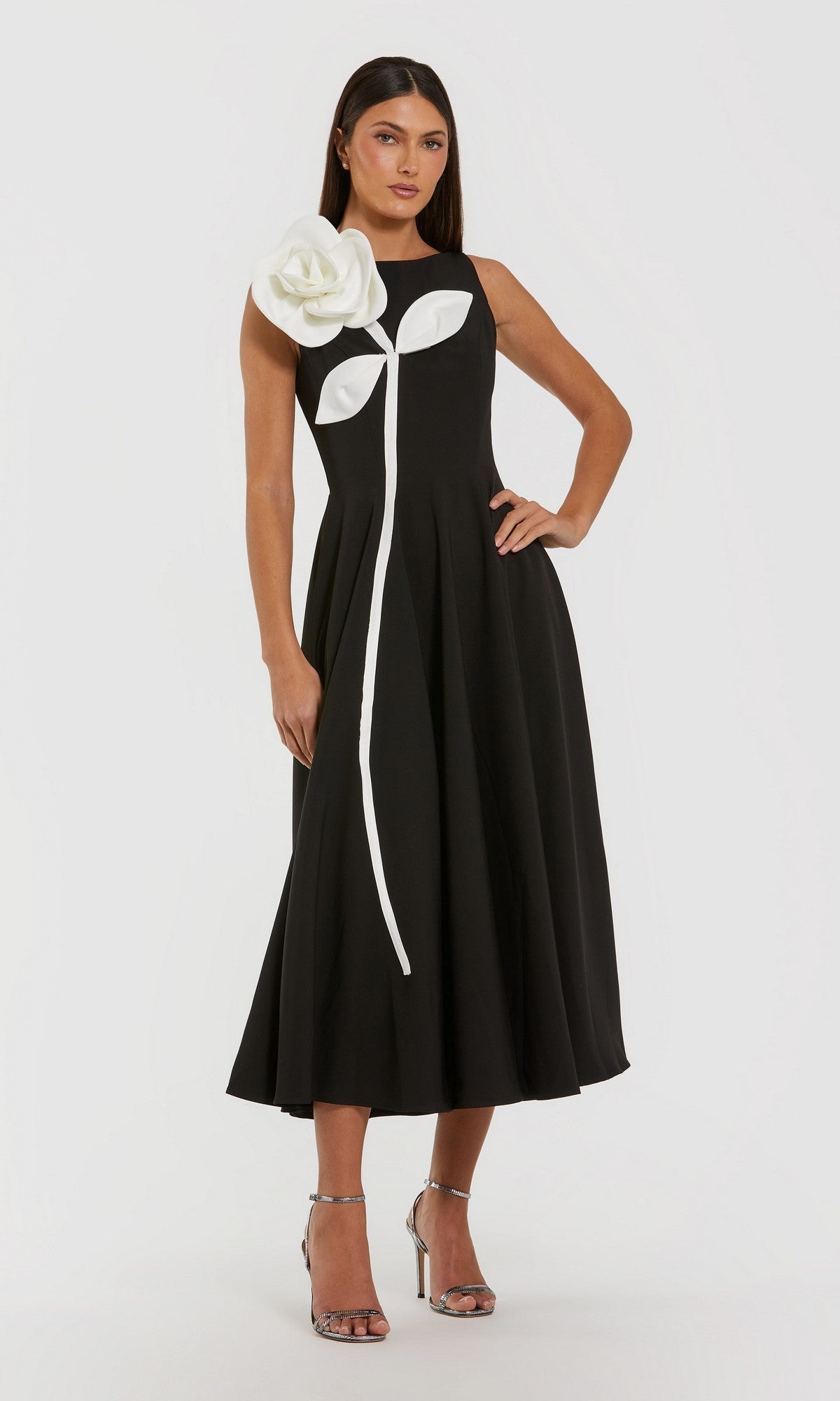 A woman stands with one hand on her hip, wearing the Mac Duggal 2329 tea-length party dress in black ivory—a sleeveless design featuring a striking 3D flower and stem detail on the bodice—paired with silver high-heeled sandals.