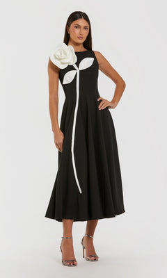 A woman stands with one hand on her hip, wearing the Mac Duggal 2329 tea-length party dress in black ivory—a sleeveless design featuring a striking 3D flower and stem detail on the bodice—paired with silver high-heeled sandals.