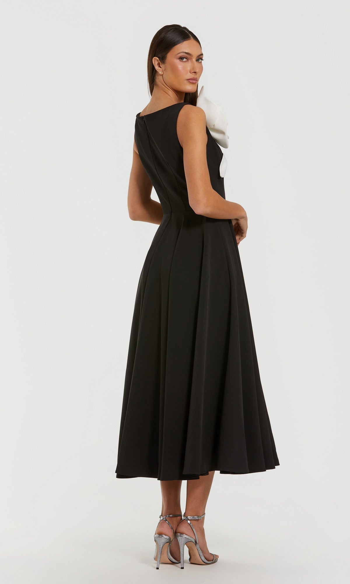 A woman poses against a plain white background wearing the Mac Duggal 2329— a sleeveless, tea-length black party dress with an elegant white ruffle detail on the front— paired with silver high-heeled sandals.