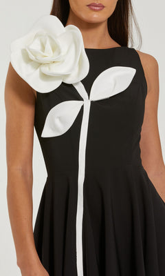 A woman wearing the Mac Duggal 2329 Tea-Length Party Dress, a sleeveless black design featuring a striking large white 3D flower with leaves detail on the shoulder and bodice.