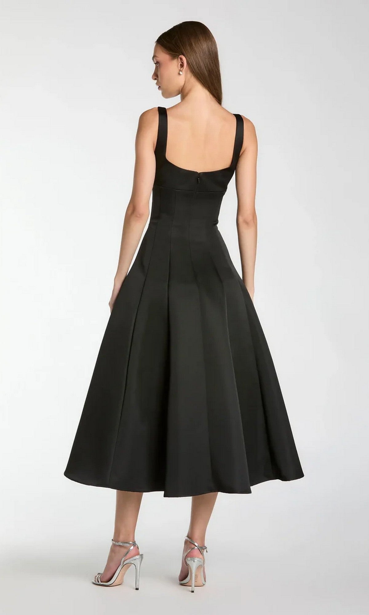 A woman stands with her back to the camera, wearing the Midi Party Dress: Mac Duggal 2331—a sleek black satin midi dress featuring a fitted bodice, flared skirt, and a delicate beaded-embellished flower detail. She pairs the elegant dress with silver high-heeled sandals, all set against a plain background.