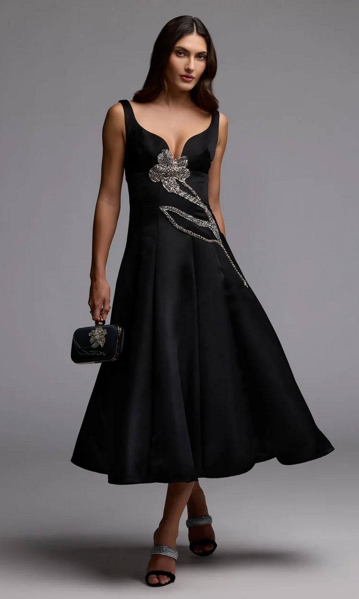 A woman stands against a plain background wearing the Mac Duggal 2331 Midi Party Dress in Black Gold, a sleeveless satin midi design adorned with beaded-embellished floral accents. She accessorizes with a small black handbag and black heeled sandals.