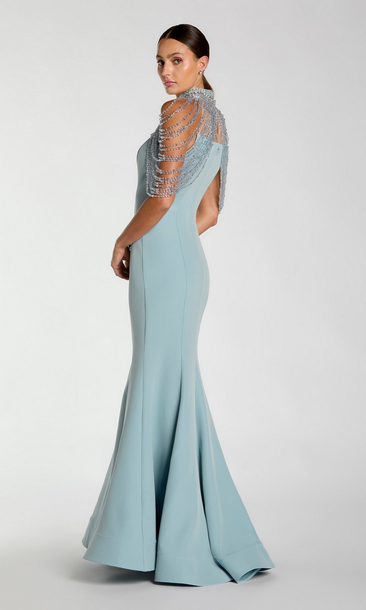 A woman in a fitted, floor-length Mac Duggal 2334 long formal dress in light blue, featuring beaded short sleeves and a mermaid silhouette, stands against a plain light background, posed slightly away from the camera.