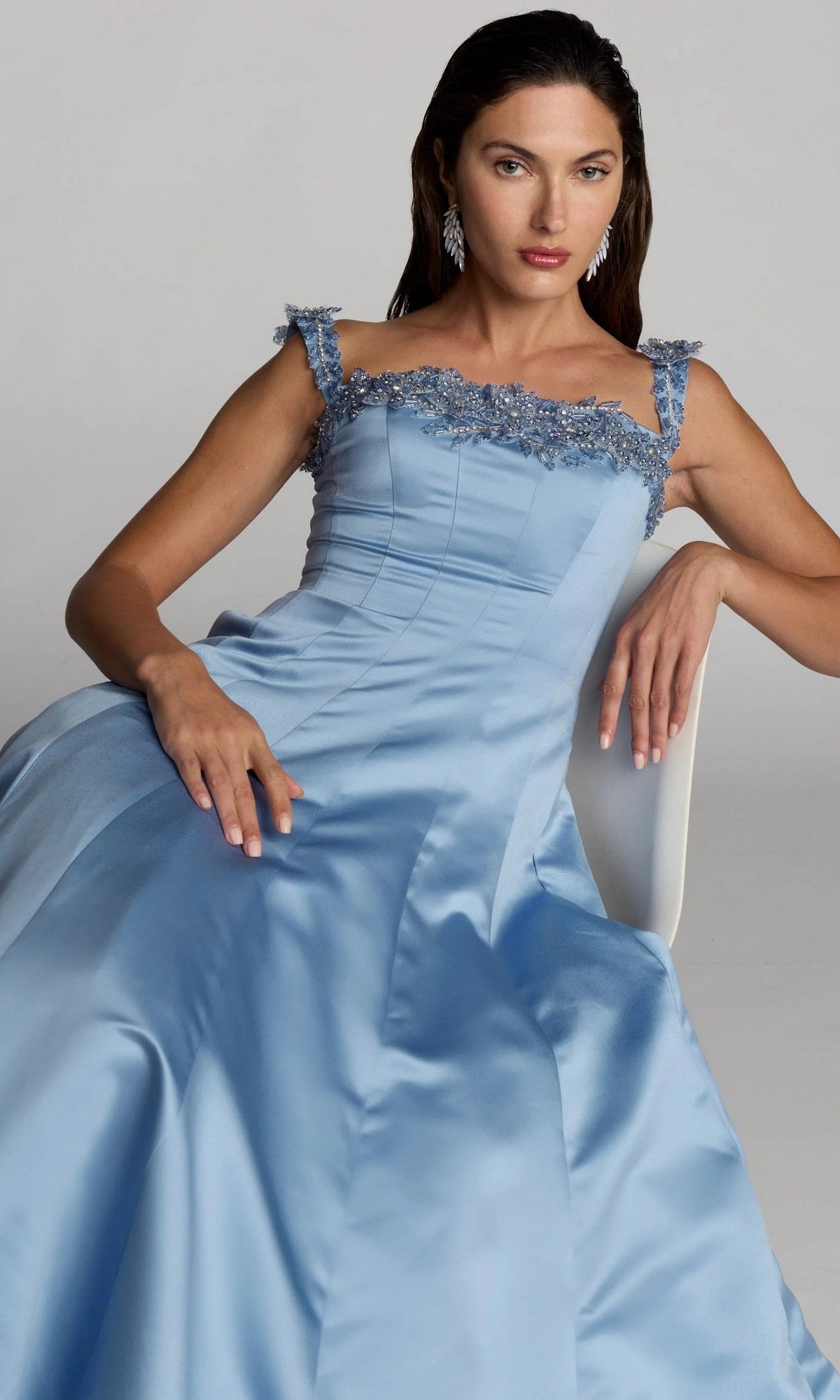 A woman sits in a white chair, looking at the camera, wearing the Mac Duggal 2337 long formal dress—a light blue satin gown featuring floral embellishments on the straps and neckline.