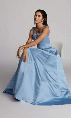 A woman wearing the Long Formal Dress: Mac Duggal 2337—a floor-length, light blue satin gown with floral details—sits on a white chair against a plain background.