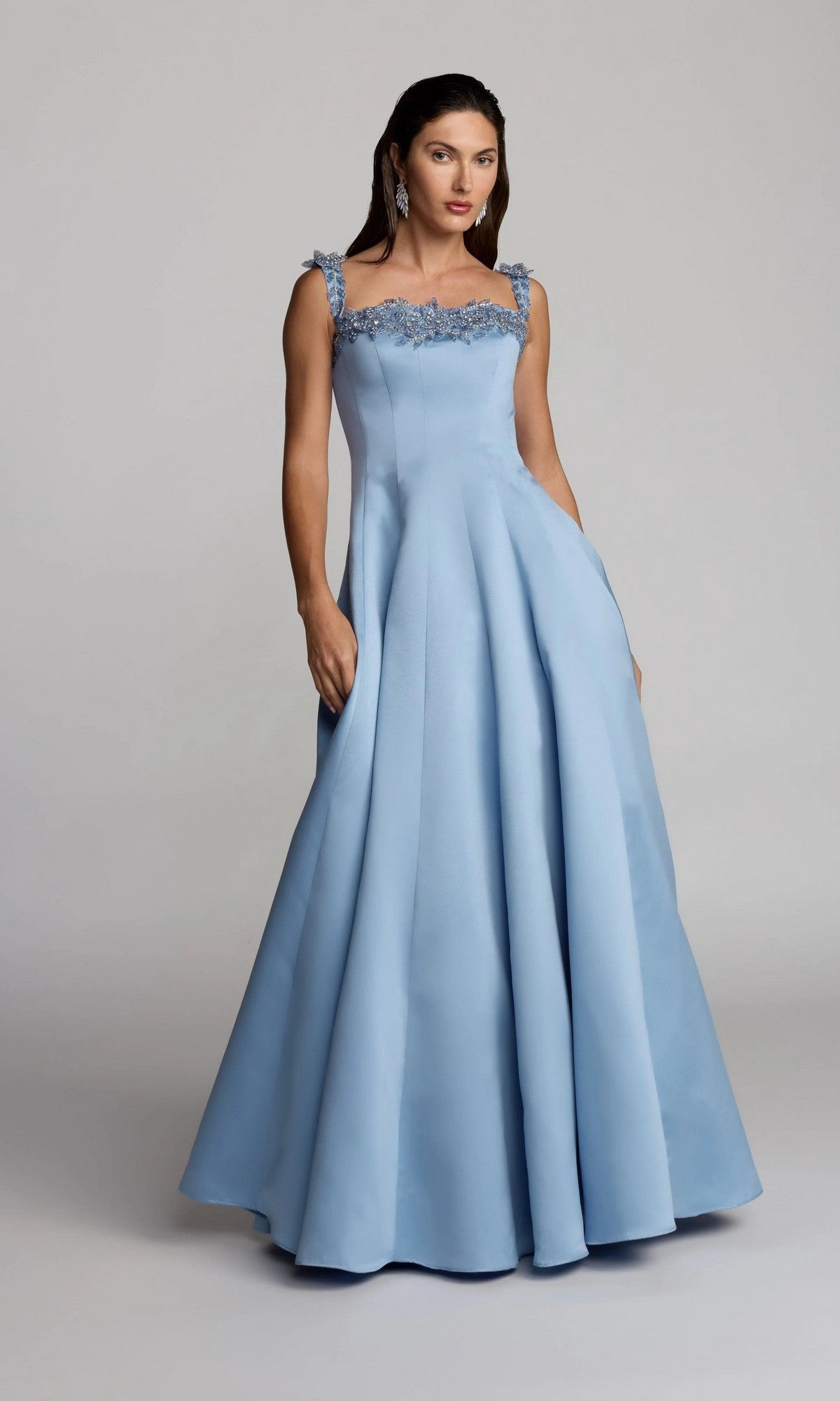 A woman wearing the Mac Duggal 2337 long formal dress in slate blue, featuring a floor-length A-line silhouette and floral strap details, stands against a plain gray background.