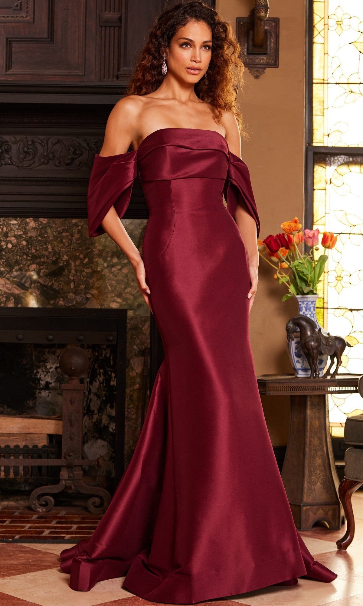 A woman wearing the Jovani 23398 Long Formal Dress—a strapless, floor-length satin and silk gown in burgundy—stands indoors near a fireplace and a table adorned with flowers and elephant figurines.