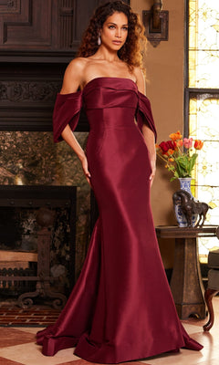 A woman wearing the Jovani 23398 Long Formal Dress—a strapless, floor-length satin and silk gown in burgundy—stands indoors near a fireplace and a table adorned with flowers and elephant figurines.