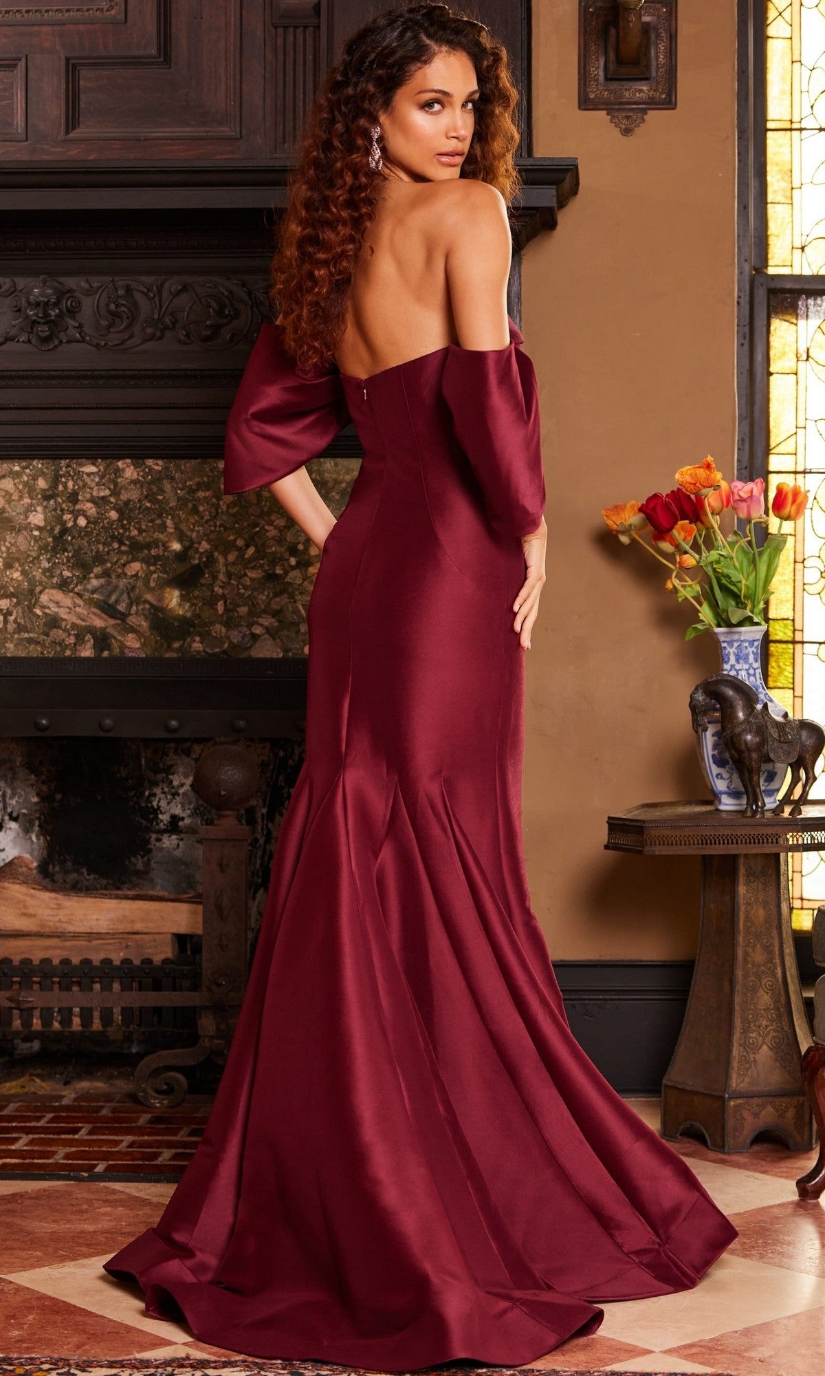 A woman wearing the elegant Jovani 23398 long formal dress—a floor-length, off-shoulder gown crafted from rich burgundy satin and silk—stands before a fireplace, glancing over her shoulder. Beside her is a table adorned with flowers and an elephant sculpture.