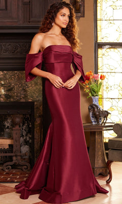 A woman stands indoors near a fireplace and a side table adorned with a flower vase and decorative elephant statues, wearing the Jovani 23398 long formal dress in elegant, off-the-shoulder satin and silk in burgundy.