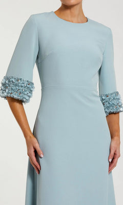 A woman is wearing the Mac Duggal 2342 long formal dress in light blue, featuring modest elbow-length sleeves, elegant beaded cuffs, and 3D floral embellishments.