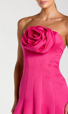 A woman wearing the Mac Duggal 2357, a long formal strapless, fitted hot pink crepe-satin dress featuring a large, sculpted fabric rose detail on the chest.