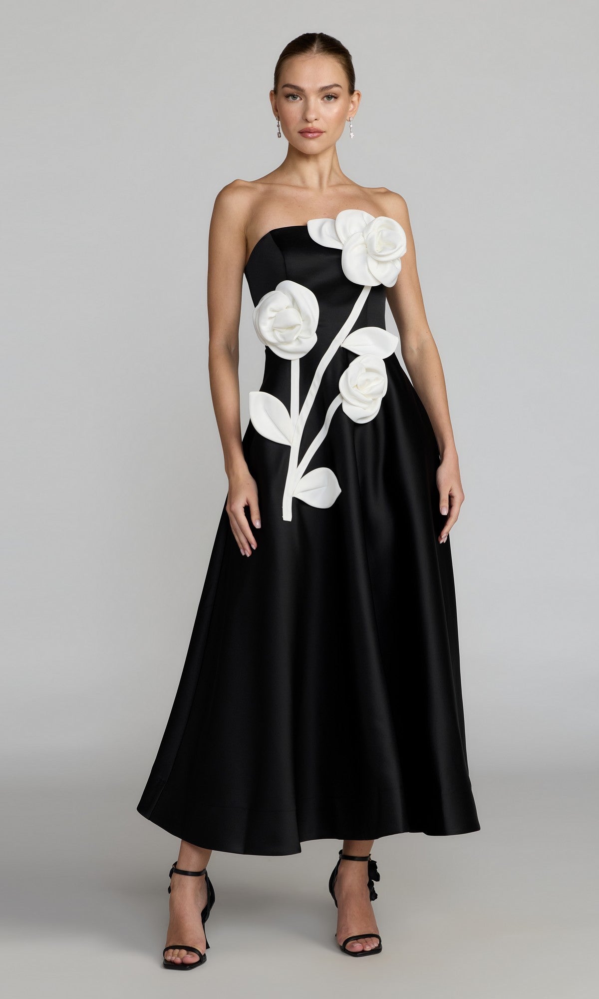 A woman stands against a plain light background, wearing the Mac Duggal 2362 long formal dress in Black Ivory—a striking black satin strapless gown embellished with large white 3D satin flowers and leaves on the front—paired with black heels.