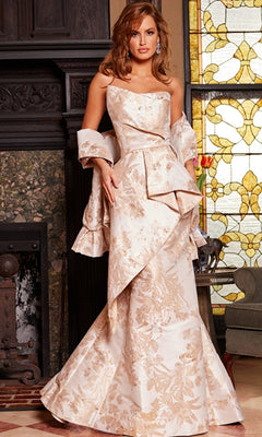 A woman in the Jovani 23624 Long Formal Dress, featuring an ornate cream and gold strapless design, stands indoors near a fireplace and stained glass window.