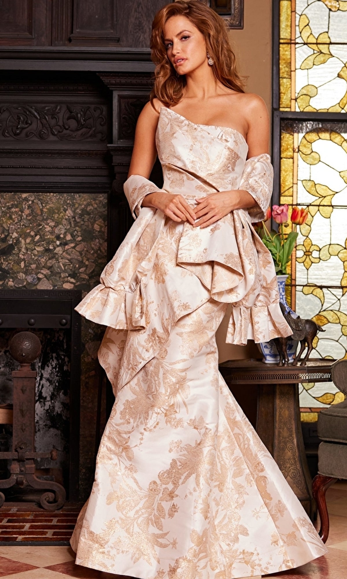 A woman stands indoors by a stained glass window and flower-topped table, wearing the Jovani 23624 Long Formal Dress—a cream, strapless gown with floral patterns, ruffles, and a matching shawl.