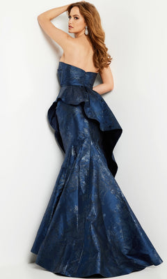 Woman wearing the Jovani 23624 Long Formal Dress, a strapless dark blue floor-length gown with peplum detail, posing with one arm raised and looking over her shoulder.
